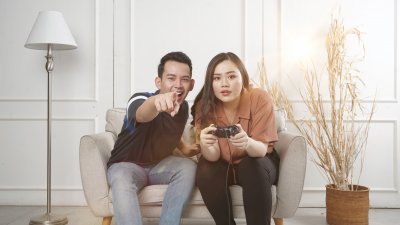 A photo of an Asian man and woman sitting on a small sofa playing video games to represent the topic of the article - Is Gaming Good For Your Mental Health And Wellbeing?