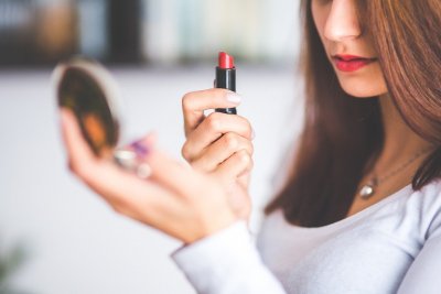 A photo of a white woman looking into a compact mirror to apply makeup and the red lipstick she's holding to represent the article title - Do I Have BDD? Making Enemies Out Of Mirrors