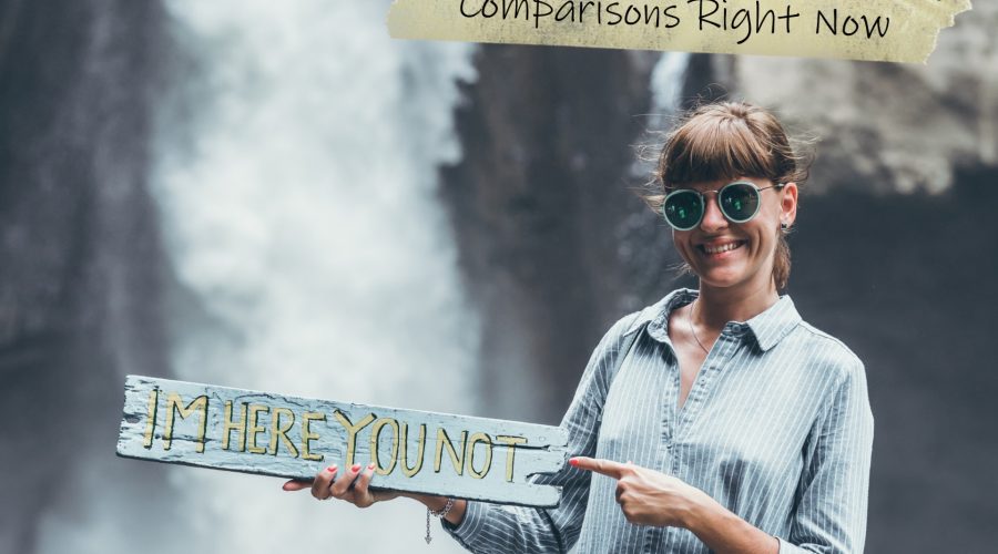 A photo of a White woman standing in front of a waterfall holding a sign that says "I'm here you not" to represent the topic of the article - Why You Need To Stop Making Comparisons Right Now