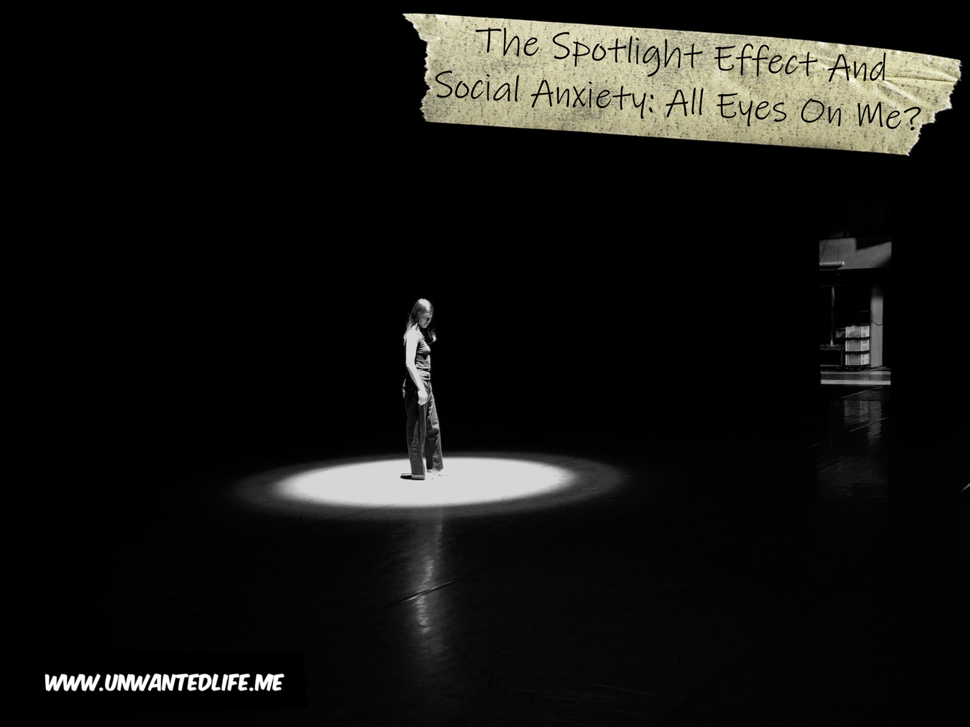 A black-and-white photo of a woman standing alone in a dark room, illuminated by a spotlight to represent the topic of the article - The Spotlight Effect And Social Anxiety: All Eyes On Me?
